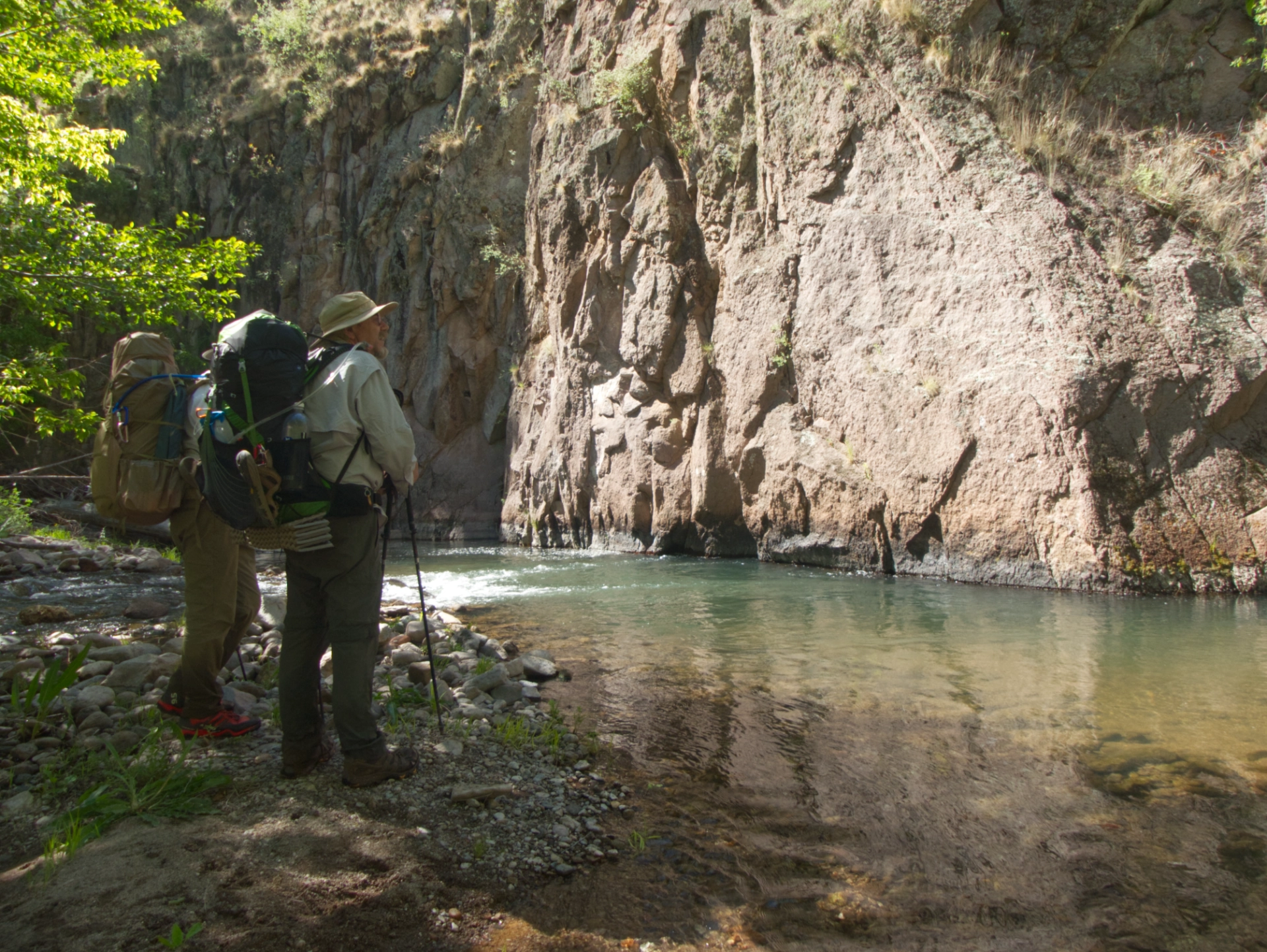 two hikers by rapids at the base of a cliff